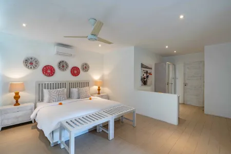 Luxurious minimalist bedroom with white king bed, orange-accented pillows, decorative wall plates, ceiling fan, and wooden bench on light wood floors. - Bali Villas